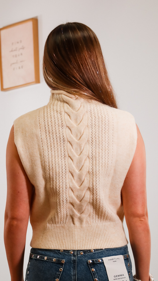 Person wearing a beige knitted vest with a braided pattern on the back, standing against a plain wall.